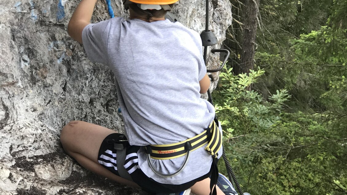 Ein Junge klettert an einer Felswand und trägt einen Sicherheitshelm. Im Hintergrund sind weitere Kletterer und Bäume zu sehen. | © Tourismusverband Schladming-Dachstein