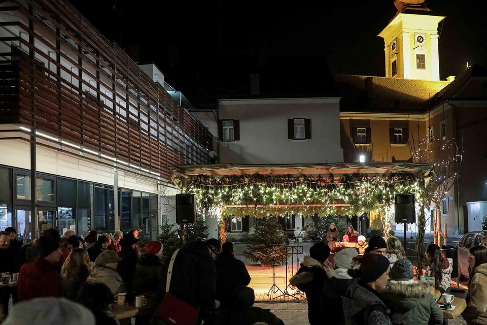 Eine festliche Veranstaltung im Freien mit einer Bühne, die mit Lichtern dekoriert ist. Im Hintergrund ist ein Kirchturm und viele Menschen sitzen und genießen die Atmosphäre. | © Foto de Monte Mureck