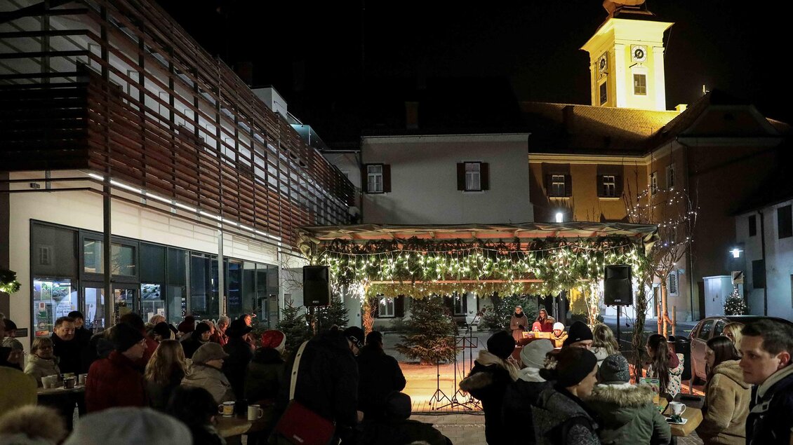 Eine festliche Veranstaltung im Freien mit einer Bühne, die mit Lichtern dekoriert ist. Im Hintergrund ist ein Kirchturm und viele Menschen sitzen und genießen die Atmosphäre. | © Foto de Monte Mureck