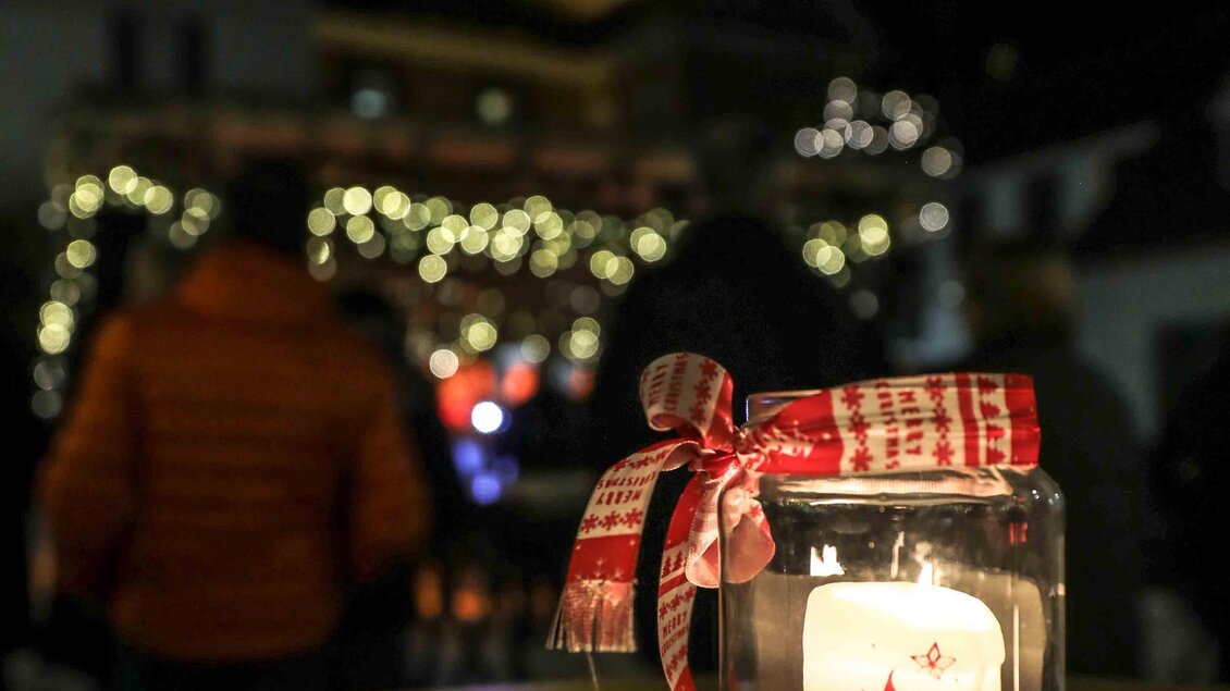 Eine festliche Kerze in einem Glas steht auf einem Tisch. Im Hintergrund sind Menschen und Lichter zu sehen, die eine winterliche Atmosphäre schaffen. | © Foto de Monte Mureck