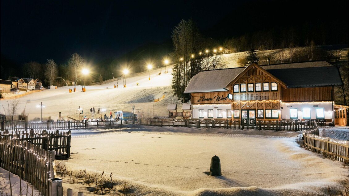 Eine charmante Berghütte in einer verschneiten Landschaft bei Nacht. Die Piste ist beleuchtet und zieht Skifahrer an. | © www.zloam.at