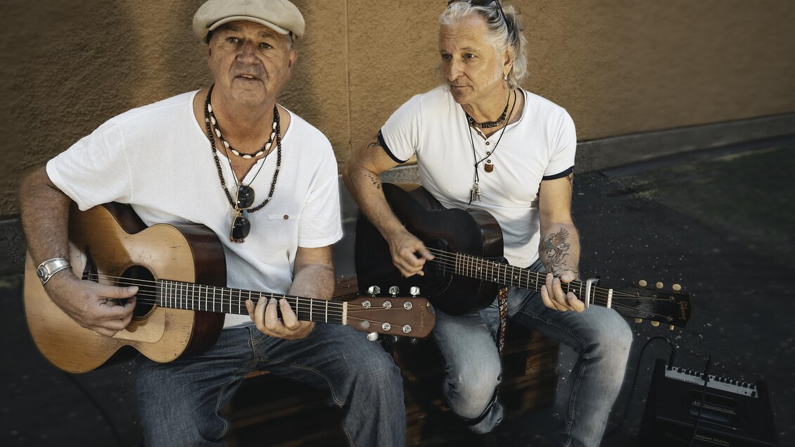 Zwei Musiker sitzen auf einer Kiste und spielen Akustikgitarre. Sie tragen lockere Kleidung und haben eine entspannte Ausstrahlung. | © Violeta Lenz