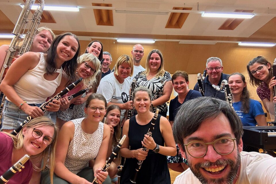 Eine Gruppe von Musikern posiert fröhlich für ein Foto in einem Proberaum. Sie halten verschiedene Blasinstrumente in den Händen und lächeln in die Kamera.