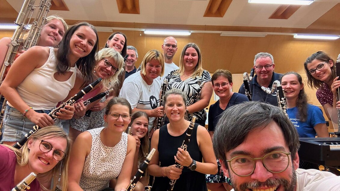 Eine Gruppe von Musikern posiert fröhlich für ein Foto in einem Proberaum. Sie halten verschiedene Blasinstrumente in den Händen und lächeln in die Kamera.