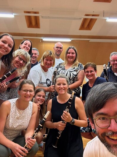 Eine Gruppe von Musikern posiert fröhlich für ein Foto in einem Proberaum. Sie halten verschiedene Blasinstrumente in den Händen und lächeln in die Kamera.