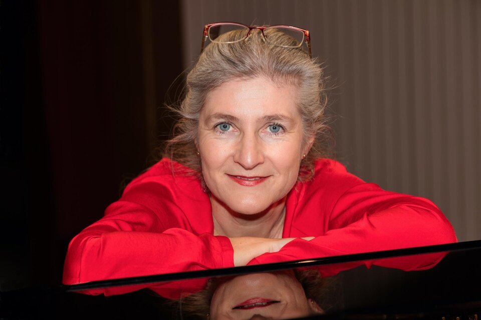 **Porträt einer Pianistin:**
Das Foto zeigt eine sympathisch Elisabeth Väth-Schadler aus Lödersdorf in einer roten Bluse, die sich entspannt auf einen glänzenden Konzertflügel stützt. Ihr direkter Blick in die Kamera und die warmen Lichtverhältnisse verleihen dem Porträt eine persönliche und elegante Ausstrahlung. Die Spiegelung im Klavier sowie die Lesebrille auf dem Kopf unterstreichen den künstlerischen Charakter der Aufnahme und vermitteln die Atmosphäre einer erfahrenen Musikerin. | © Elisabeth Väth-Schadler