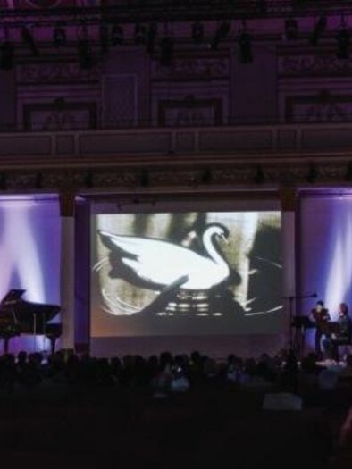 Ein Konzertsaal mit einem Klavier, auf dem ein Musiker spielt. Im Hintergrund wird ein Projektorbild mit einem Schwan gezeigt, umgeben von sanften Lichtern. | © Oststeiermark Tourismus