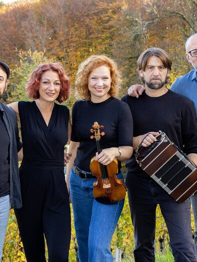 Eine Gruppe von fünf Musikern steht in einer malerischen Landschaft mit buntem Laub im Hintergrund. Sie halten verschiedene Musikinstrumente und lächeln in die Kamera. | © Harry Schiffer