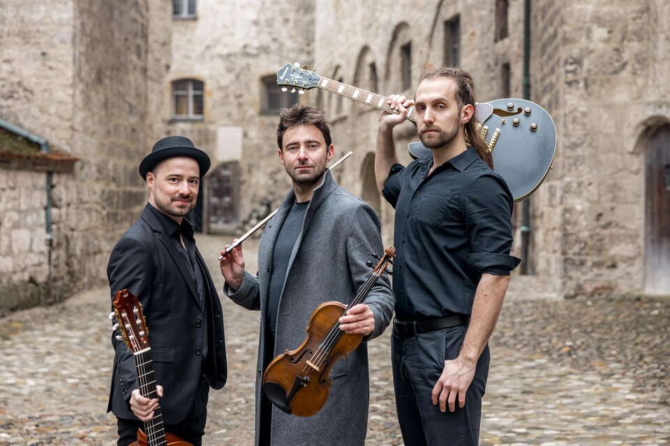 3 dunkel gekleidete Musiker mit einer akustischen Gitarre, einer Geige und einer E-Gitarre stehen repräsentativ vor einerm altertümlichen Hintergrund einer Burg. | © Oststeiermark Tourismus