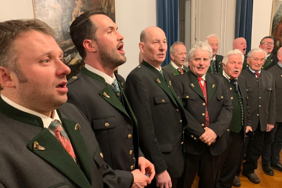 Eine Gruppe von Sängern in traditionellen Trachten singt gemeinsam in einem festlichen Raum. Im Hintergrund sind Kunstwerke und Vorhänge sichtbar. | © Männergesangsverein Admont