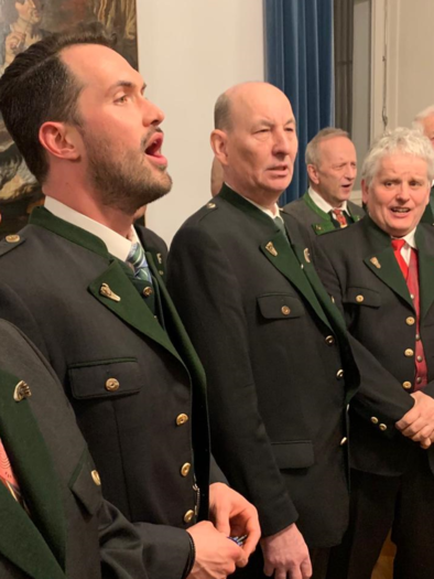 A group of singers in traditional costumes sings together in a festive space. In the background, artworks and curtains are visible. | © Männergesangsverein Admont