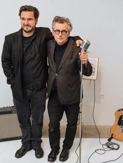 Zwei Männer stehen nebeneinander und lächeln. Im Hintergrund sind ein Mikrofon, ein Gitarrenverstärker und eine Gitarre zu sehen. | ©  David Višnjić