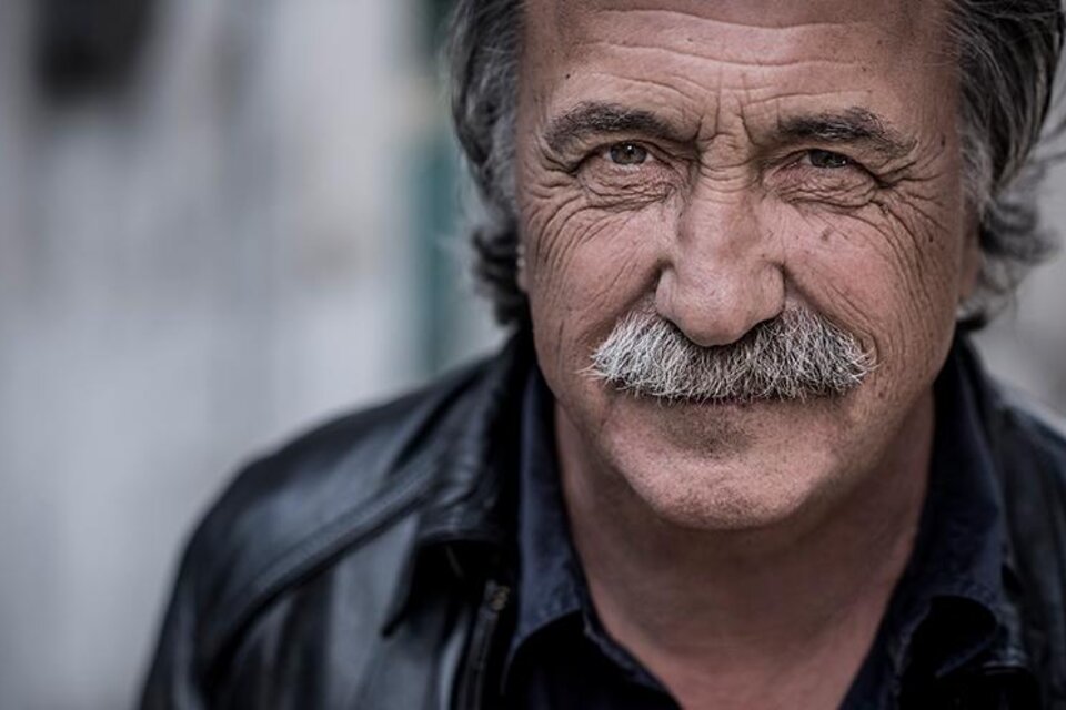 Ein älterer Mann mit einem markanten Schnurrbart und einem freundlichen Lächeln. Er trägt eine schwarze Lederjacke und hat graue Haare.