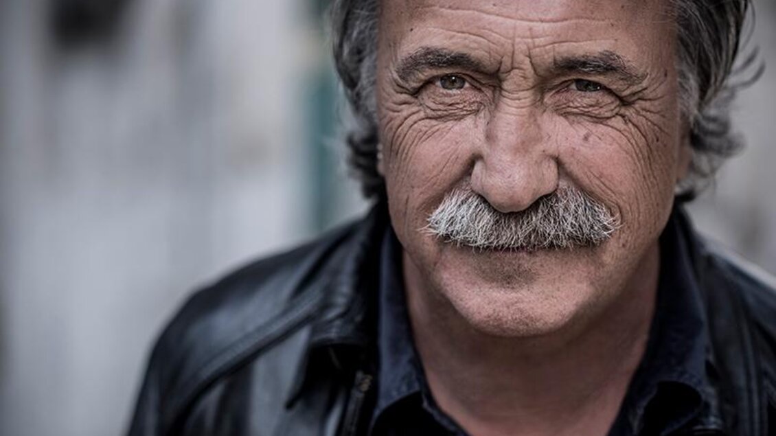 Ein älterer Mann mit einem markanten Schnurrbart und einem freundlichen Lächeln. Er trägt eine schwarze Lederjacke und hat graue Haare.