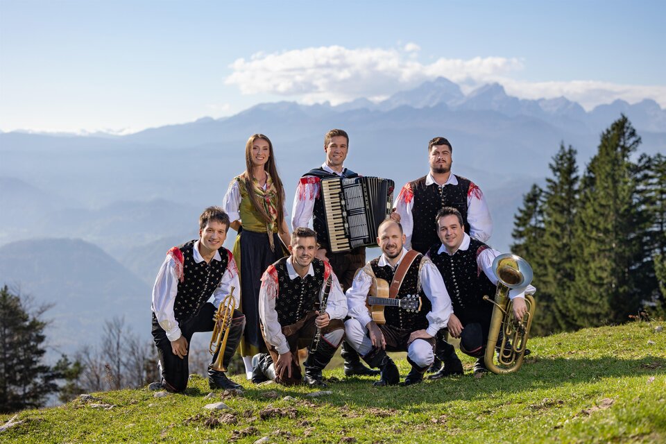 Eine Gruppe von sieben Musikern posiert auf einer Wiese mit Bergen im Hintergrund. Sie tragen traditionelle Trachten und halten verschiedene Instrumente.