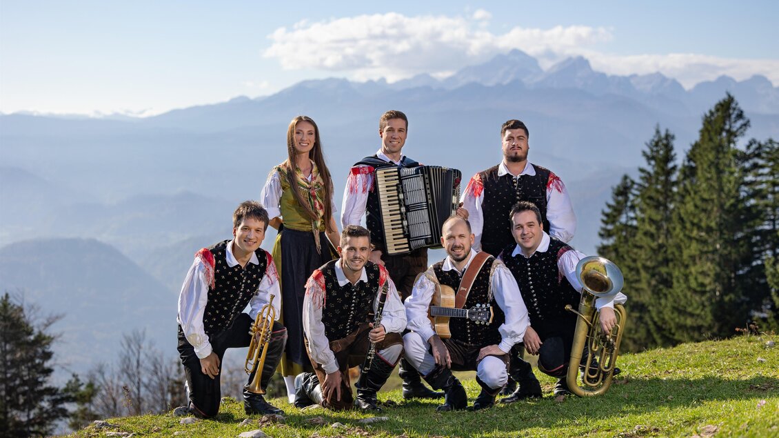 Eine Gruppe von sieben Musikern posiert auf einer Wiese mit Bergen im Hintergrund. Sie tragen traditionelle Trachten und halten verschiedene Instrumente.