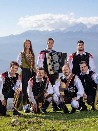 Eine Gruppe von sieben Musikern posiert auf einer Wiese mit Bergen im Hintergrund. Sie tragen traditionelle Trachten und halten verschiedene Instrumente.