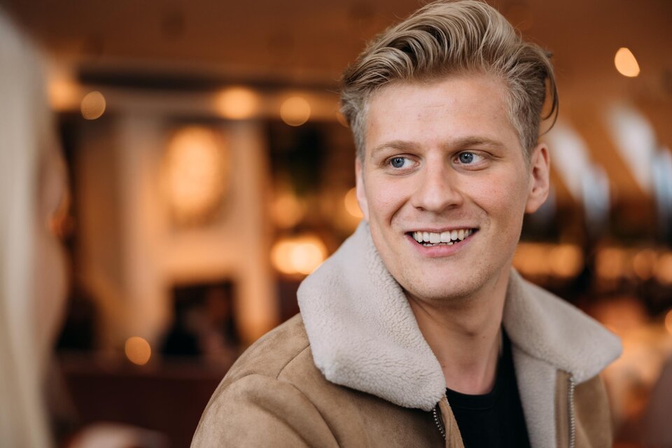 Ein lächelnder junger Mann mit blonden Haaren trägt eine braune Jacke mit einem weißen Kragen. Er scheint in einem gemütlichen Café zu sein.