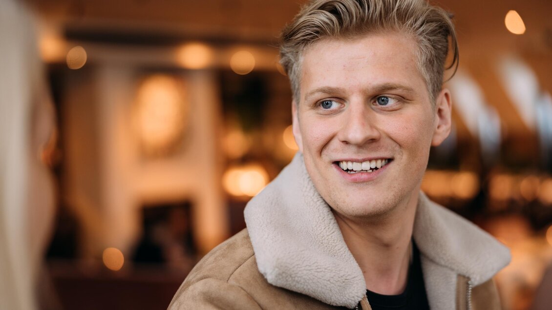 Ein lächelnder junger Mann mit blonden Haaren trägt eine braune Jacke mit einem weißen Kragen. Er scheint in einem gemütlichen Café zu sein.