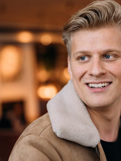 Ein lächelnder junger Mann mit blonden Haaren trägt eine braune Jacke mit einem weißen Kragen. Er scheint in einem gemütlichen Café zu sein.