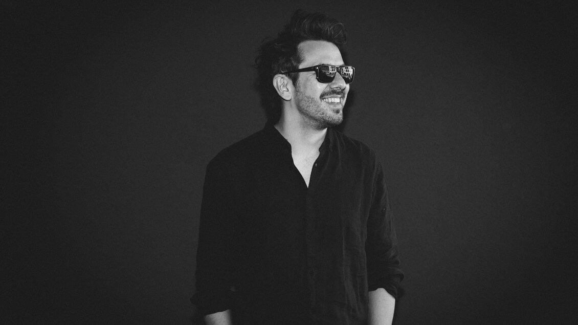 Ein lächelnder Mann trägt eine schwarze Sonnenbrille und ein schwarzes Hemd. Der Hintergrund ist dunkel und verleiht dem Bild eine stilvolle Atmosphäre.