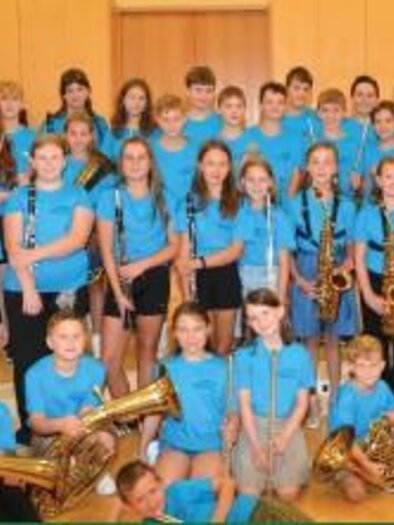 Gruppenfoto einer Kinder- und Jugendblasmusik: Viele Kinder und Jugendliche in blauen T-Shirts stehen und sitzen in mehreren Reihen in einem Saal und halten verschiedene Blasinstrumente wie Saxofone, Klarinetten, Hörner und eine Tuba. | © Marktmusikkapelle Irdning