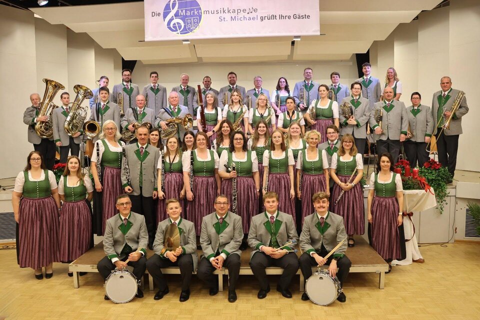 Eine Gruppe von Menschen in traditionellen Kleidungen pose für ein Foto. Sie stehen und sitzen in einem hellen Raum mit rustikalem Hintergrund. | © Marktmusikkapelle St. Michael