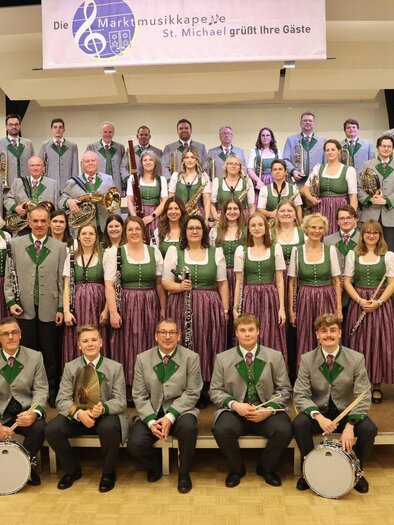 Eine Gruppe von Menschen in traditionellen Kleidungen pose für ein Foto. Sie stehen und sitzen in einem hellen Raum mit rustikalem Hintergrund. | © Marktmusikkapelle St. Michael