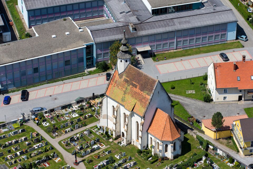Eine Kirche mit einem roten Satteldach steht neben einem Friedhof. Im Hintergrund sind moderne Gebäude zu sehen.