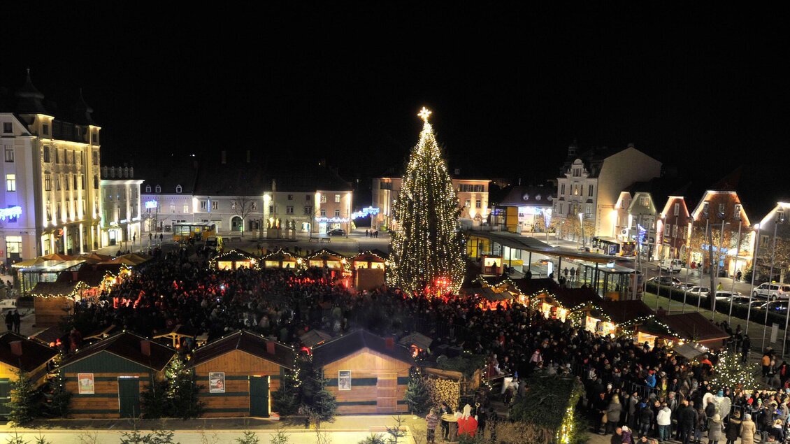 Ein festlicher Weihnachtsmarkt bei Nacht mit vielen Menschen und bunten Lichtern. In der Mitte steht ein großer, beleuchteter Weihnachtsbaum. | © Stadt Bruck/Mur, M.Maili