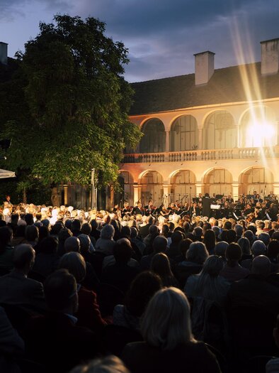 Ein Konzert im Freien bei Abenddämmerung. Die Zuschauer sind vor einem historischen Gebäude versammelt, während die Bühne beleuchtet ist. | © Bernhard Bergmann
