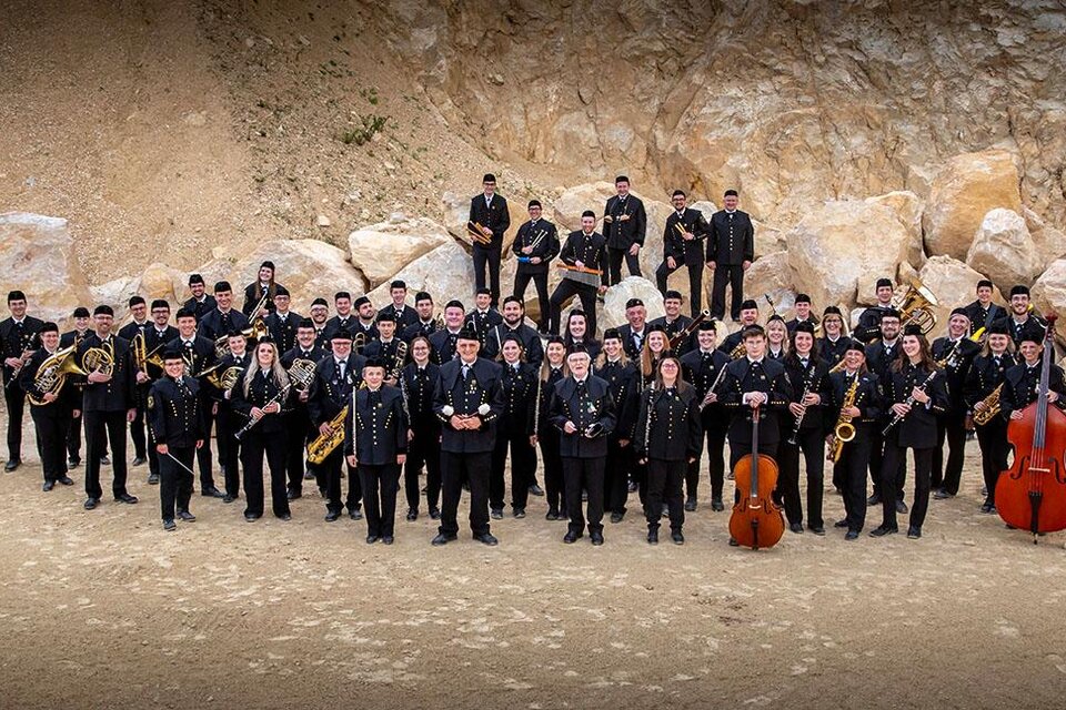 Eine große Musikkapelle steht vor einer felsigen Kulisse. Die Mitglieder tragen schwarze Kleidung und halten verschiedene Musikinstrumente. | © August Zoebl