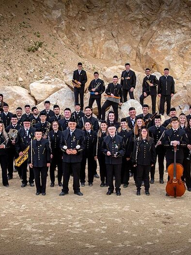 A large marching band stands in front of a rocky backdrop. The members wear black clothing and hold various musical instruments. | © August Zoebl