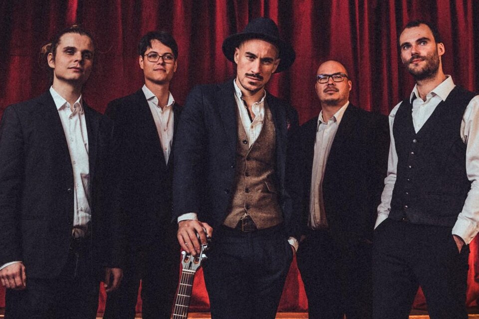 Eine Band aus fünf Männern steht vor einem roten Vorhang. Sie tragen elegante Anzüge und schauen freundlich in die Kamera.