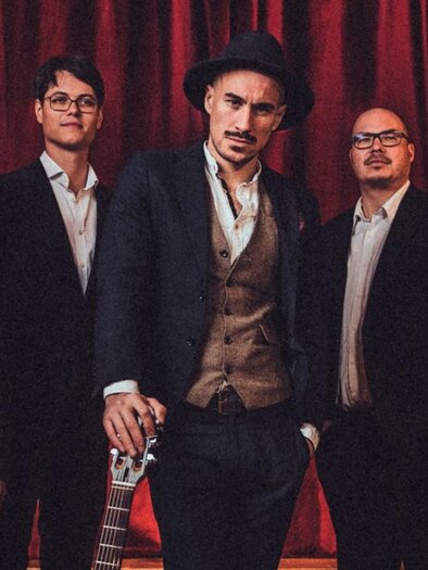 A band of five men stands in front of a red curtain. They are wearing elegant suits and are looking friendly at the camera.