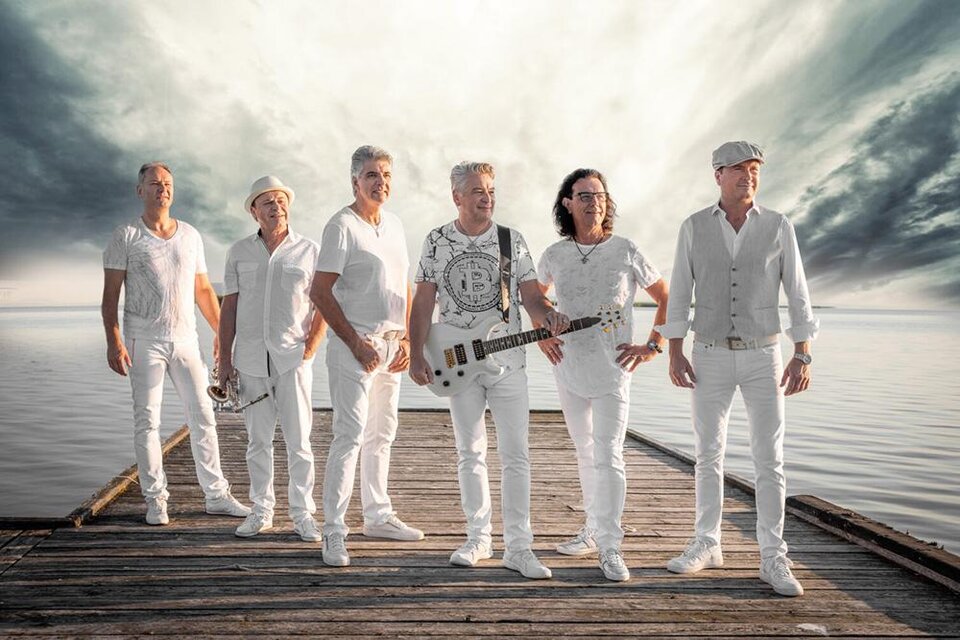 Eine Band in weißen Outfits steht auf einem Steg am Wasser. Der Himmel ist dramatisch und bewölkt, was eine dynamische Atmosphäre schafft. | © Karl Schroter