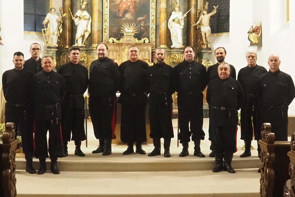 Eine Gruppe von Männern in dunkler Kleidung steht in einer Kirche vor einem kunstvoll gestalteten Altar. Sie blicken in die Kamera und scheinen bereit für ein Ereignis oder eine Aufführung zu sein. | © DKCWH
