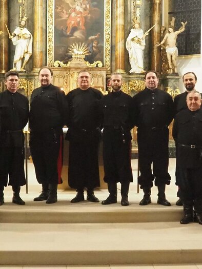 Eine Gruppe von Männern in dunkler Kleidung steht in einer Kirche vor einem kunstvoll gestalteten Altar. Sie blicken in die Kamera und scheinen bereit für ein Ereignis oder eine Aufführung zu sein. | © DKCWH