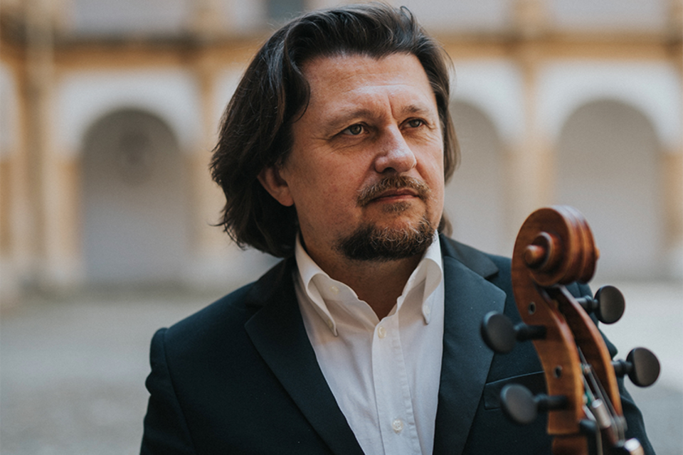 Ein Musiker in einem eleganten Anzug steht mit einem Cello. Der Hintergrund zeigt eine historische Architektur. | © Lukas Elsneg