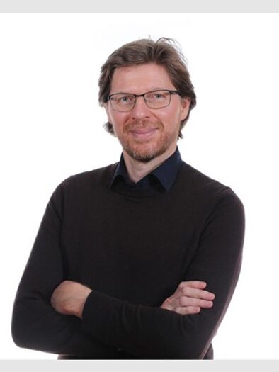 Ein Mann mit Brille steht lächelnd vor einem weißen Hintergrund. Er trägt einen schwarzen Pullover und hat die Arme verschränkt. | © Bild: Foto Freisinger