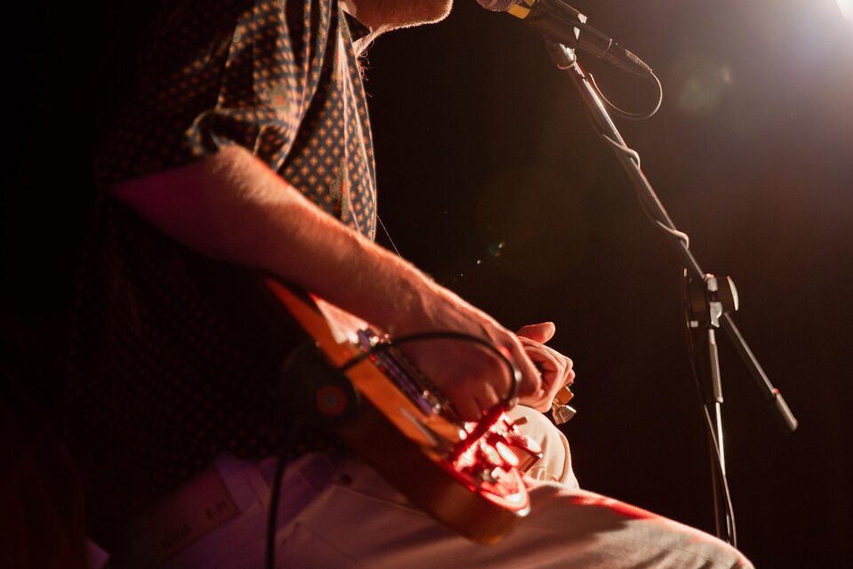 Ein Musiker sitzt auf einem Stuhl und spielt Gitarre. Er ist im Spotlight und scheint sich auf seine Darbietung zu konzentrieren. | © TVB Südsteiermark – Achromatic Photography