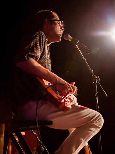 Ein Musiker sitzt auf einem Stuhl und spielt Gitarre. Er ist im Spotlight und scheint sich auf seine Darbietung zu konzentrieren. | © TVB Südsteiermark – Achromatic Photography