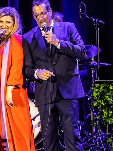 A pair of singers stands on stage and speaks into the microphone. In the background, musical instruments and dim stage lighting can be seen. | © (c)Andreas Müller