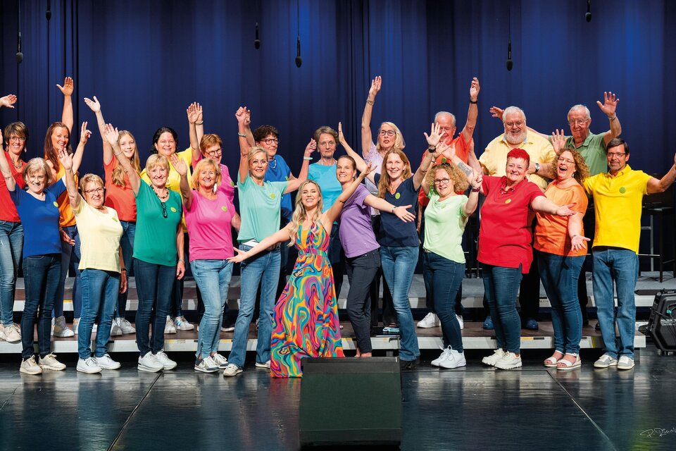 Eine fröhliche Gruppe von Sängern und Sängerinnen steht auf der Bühne und posiert zusammen. Sie tragen bunte T-Shirts und zeigen ihre Energie und Freude während eines Jubiläumskonzerts. | © MG Lannach - Petru Rimovetz