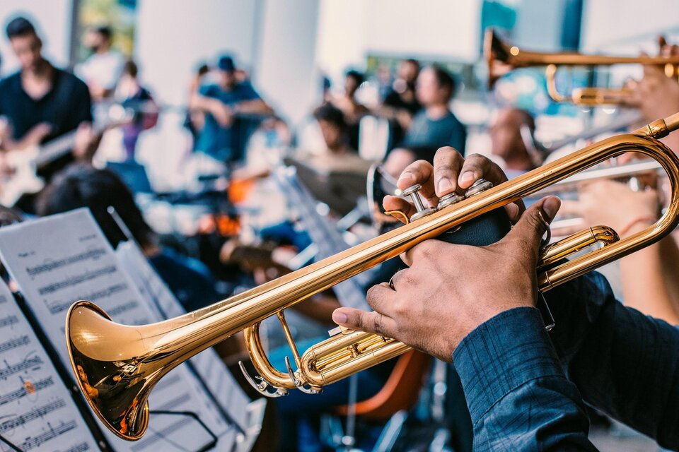 Eine Person spielt Trompete in einer musikalischen Umgebung. Im Hintergrund sind weitere Musiker und Notenblätter zu sehen. | © Unsplash