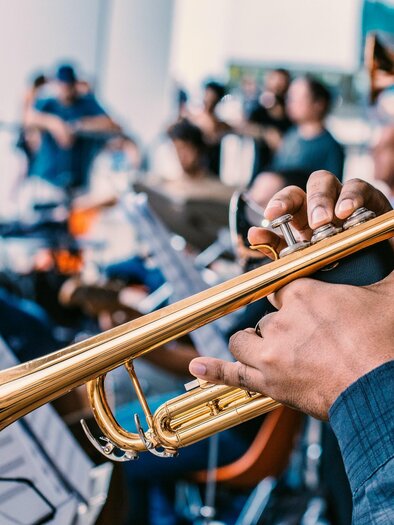 A person is playing the trumpet in a musical setting. In the background, more musicians and sheet music can be seen. | © Unsplash