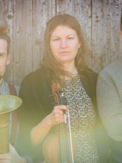 Eine Gruppe von drei Musikern posiert vor einer Holzoberfläche. Sie halten verschiedene Instrumente und blicken in die Kamera. | © Christoph Huber
