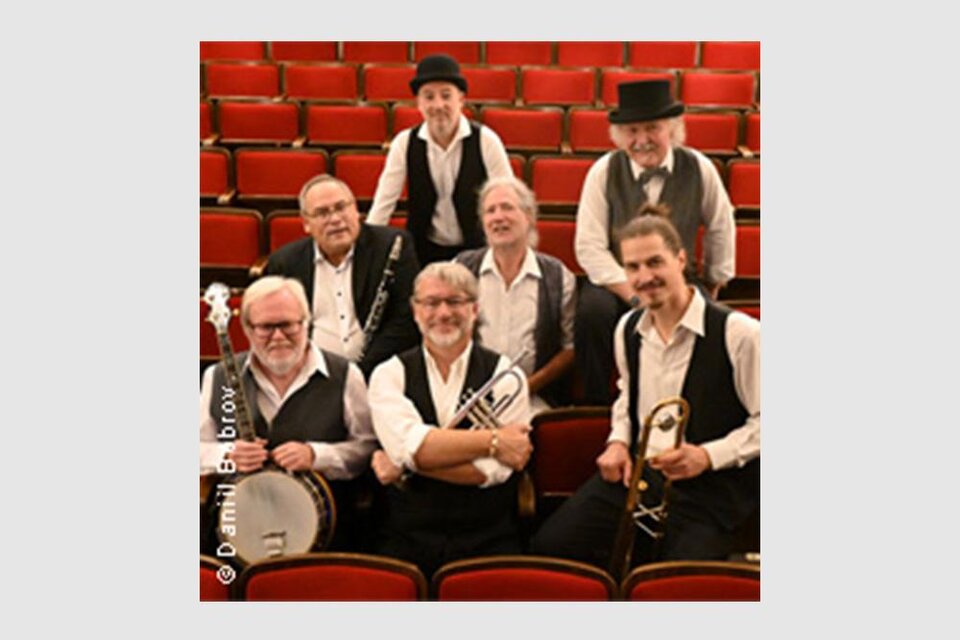 Eine Gruppe von acht Musikern in historischen Outfits sitzt in einem Theater mit roten Sitzen. Sie halten Instrumente und lächeln in die Kamera. | © Daniil Bobrov