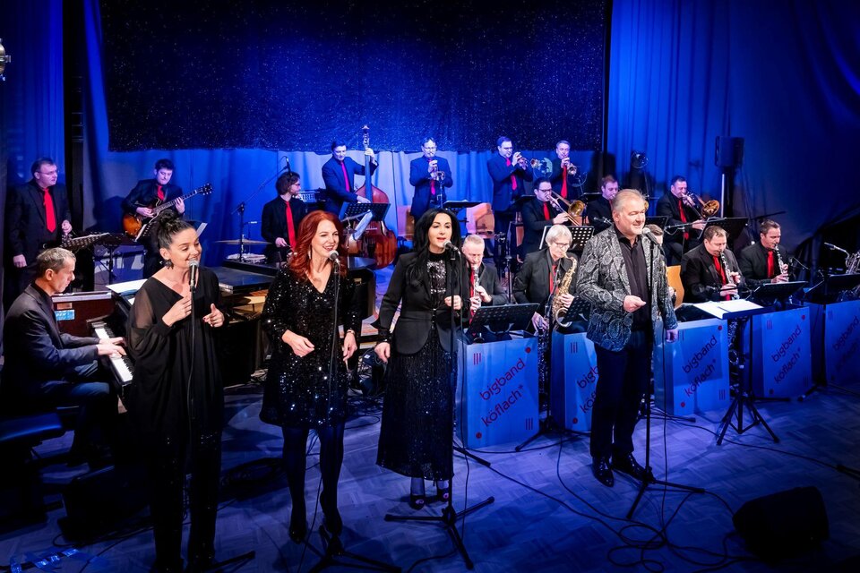 Eine lebhafte Musikaufführung mit mehreren Sängern und einer großen Band im Hintergrund. Die Künstler sind elegant gekleidet und strahlen Energie aus. | © Petru Rimovetz