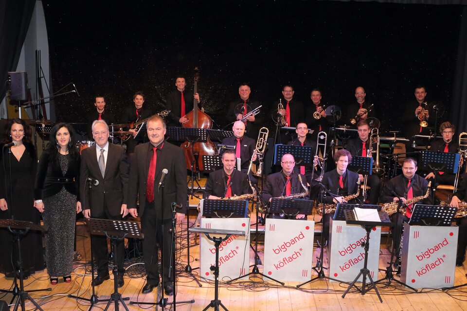 Eine große Gruppe von Musikern steht auf der Bühne, die in formeller Kleidung gekleidet sind. Im Vordergrund stehen einige Sänger, während die Band im Hintergrund spielt. | © Bigband Köflach